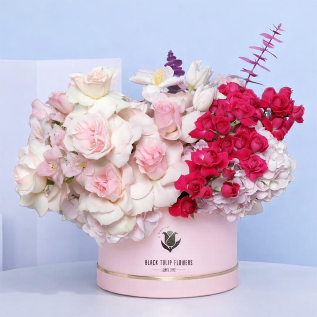 A pink floral arrangement featuring light and dark pink roses, tulips, hydrangeas, and eucalyptus in a decorative box.