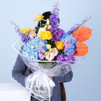 Stunning bouquet with hydrangeas, Anthurium, peach Lisianthus, roses, Mathiola, and Delphinium, elegantly wrapped in glass sheet.