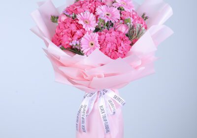 A vibrant pink bouquet featuring gerberas, hydrangeas, roses, and ferns, wrapped in pink tissue paper and tied with white ribbon
