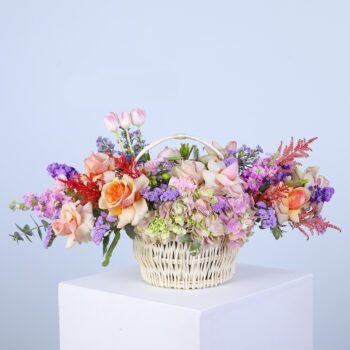 A basket filled with pastel-colored flowers, including hydrangeas, roses, carnations, and greenery.