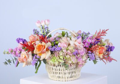 A basket filled with pastel-colored flowers, including hydrangeas, roses, carnations, and greenery.
