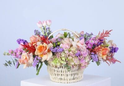 A basket filled with pastel-colored flowers, including hydrangeas, roses, carnations, and greenery.