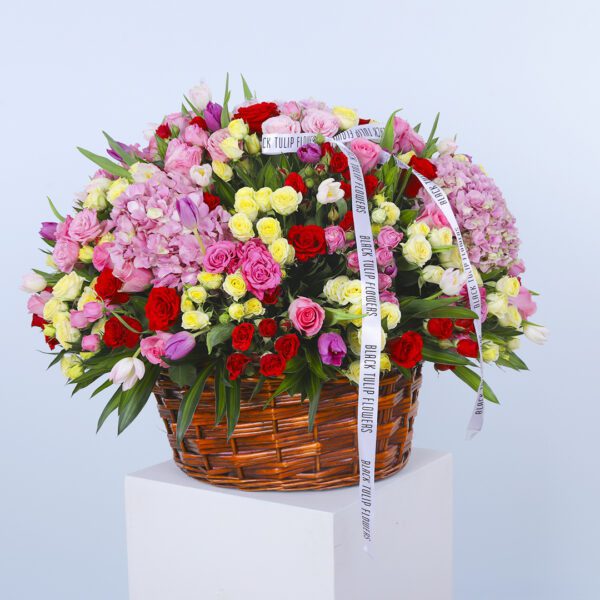 A large basket overflowing with pink hydrangeas, mixed-color roses, and greenery, tied with a ribbon.