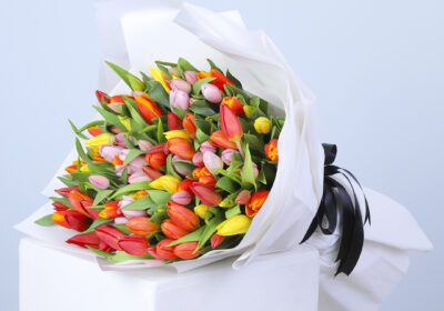A colorful bouquet of tulips in red, orange, yellow, and pink, wrapped in white paper with a black ribbon