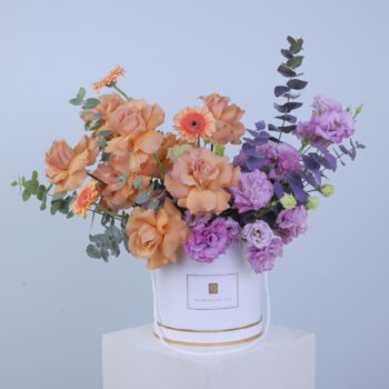 A vibrant bunch of peach roses, gerbera daisies, and purple lisianthus arranged in a white box.