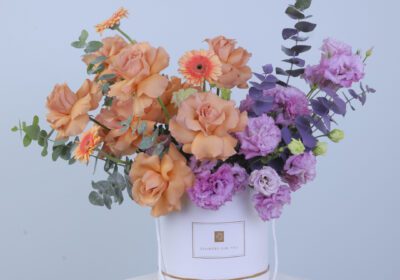 A vibrant bunch of peach roses, gerbera daisies, and purple lisianthus arranged in a white box.