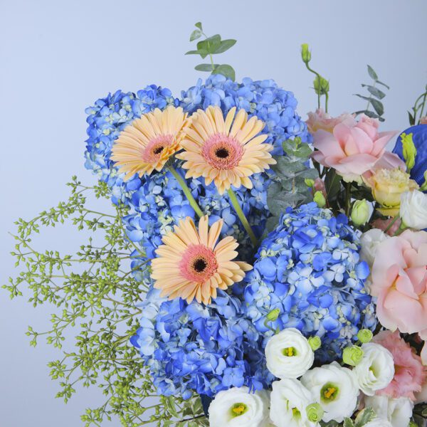 A stunningly composed arrangement of blue hydrangeas, daisies, and peach and white carnations, artfully presented in a white box with a touch of greenery.