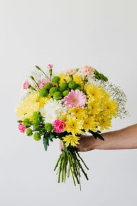 A hand holding a colorful bouquet of flowers, including yellow chrysanthemums, green button poms, pink carnations, and white gypsophila, against a white background.