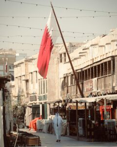 The image shows a street scene in Souq Waqif, a traditional marketplace in Doha, Qatar. A large Qatari flag is prominently displayed on a flagpole, waving in the wind. The street is lined with traditional Qatari architecture, featuring buildings with sand-colored walls and wooden balconies.