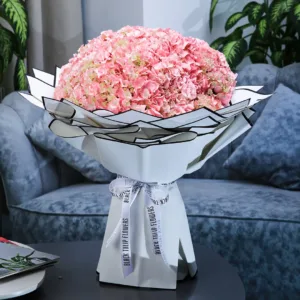 The image shows a large bouquet of pink hydrangeas. The hydrangeas are arranged in a large, round shape and wrapped in white paper with a black stripe.