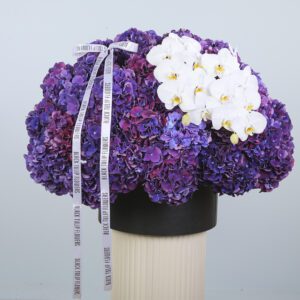 The image shows a large, round bouquet of purple hydrangeas with a cluster of white orchids on top. The bouquet is arranged in a black vase