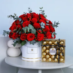 The image shows a beautiful arrangement of red roses in a white box. There is also a box of Ferrero Rocher chocolates next to it. The combination of flowers and chocolates makes it a perfect gift for a loved one.