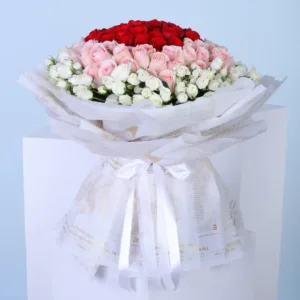 The image shows a large bouquet of roses. The bouquet is wrapped in white paper and tied with a white ribbon. The roses are arranged in a way that creates a gradient effect, with red roses at the top, pink roses in the middle, and white roses at the bottom.