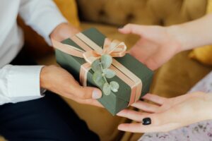 The image shows a man presenting a gift to a woman. The gift is wrapped in green paper with a pink ribbon and decorated with a sprig of eucalyptus. The woman is smiling and reaching out to take the gift.