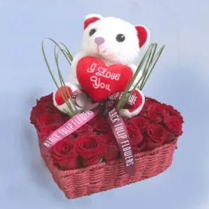 The image shows a heart-shaped basket filled with red roses. A cute teddy bear is sitting on top of the roses, holding a heart that says "I Love You." The basket is decorated with a ribbon