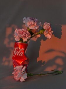 A crumpled, red Coca-Cola can sits on a gray surface, holding several white and pink flowers.