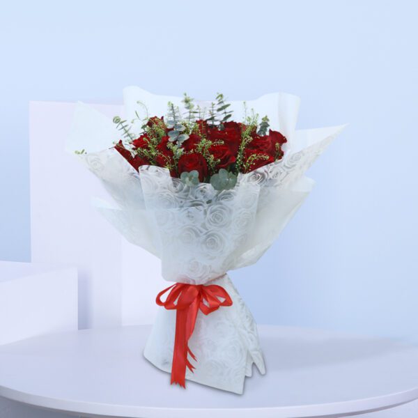 Bouquet of red roses with lush greenery, wrapped in white with a red ribbon.