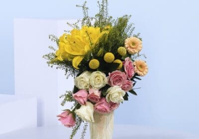 A marble vase filled with yellow lilies, pink and white roses, peach gerberas, and lush greenery.