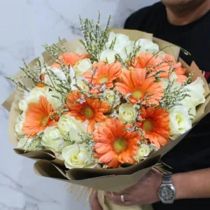 a bouquet of flowers wrapped in brown paper. The bouquet features a mix of white roses and orange gerbera daisies, accented with small white flowers with delicate branching stems