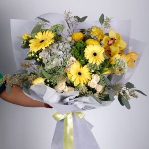 a bouquet of flowers wrapped in white paper against a light grey background. The bouquet features a mix of yellow gerbera daisies, yellow orchids, small yellow button flowers, white roses, and blue hydrangeas.