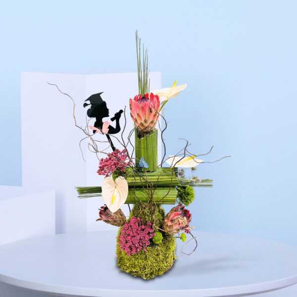 Graduation-themed flower arrangement with proteas, pink anthuriums, moss base, and a silhouette of a graduate holding a diploma, styled with natural twigs and butterflies.