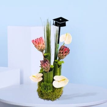 Graduation-themed floral arrangement with pink proteas, white anthuriums, tall green foliage, moss base, and a black graduation cap topper.