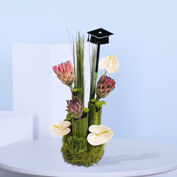 Graduation-themed floral arrangement with pink proteas, white anthuriums, tall green foliage, moss base, and a black graduation cap topper.