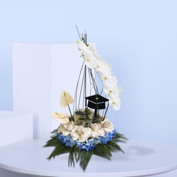 Graduation-themed floral arrangement with white orchids, blush roses, pink anthuriums, blue hydrangeas, and a black graduation cap topper.