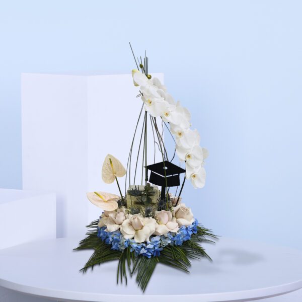 Graduation-themed floral arrangement with white orchids, blush roses, pink anthuriums, blue hydrangeas, and a black graduation cap topper.