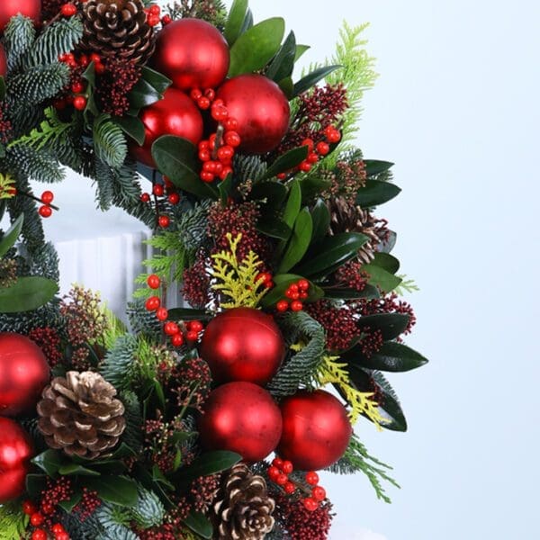 03 Christmas wreath with evergreens, red ornaments, berries, and pinecones on a white stand.
