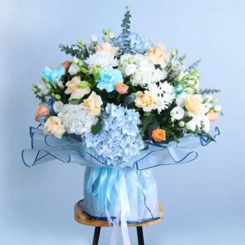 Bouquet featuring hand-tied blue hydrangeas, cream roses, lisianthus & chrysanthemums in blue wrap, finished with layered ribbons.
