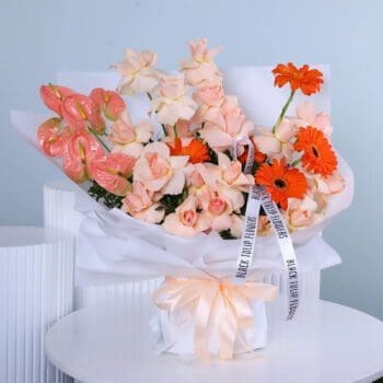 A bouquet with peach roses, orange gerberas, peach anthurium, and greenery in elegant white wrapping with a peach ribbon.