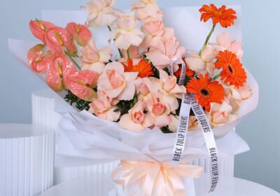A bouquet with peach roses, orange gerberas, peach anthurium, and greenery in elegant white wrapping with a peach ribbon.