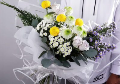 Bouquet featuring hand-tied yellow ping pongs, green ping pongs & purple roses in clear wrap, finished with greenery.