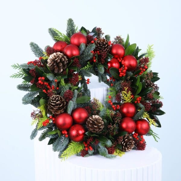 red-holiday-wreath-christmas-decor Christmas wreath with evergreens, red ornaments, berries, and pinecones on a white stand.