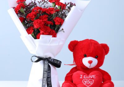 Red carnations with wax flowers in white wrap, paired with a love teddy