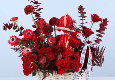 Red roses, red anthurium, red gerbera daisies, and red foliage in a wicker basket arrangement style, finished with a printed ribbon bow.