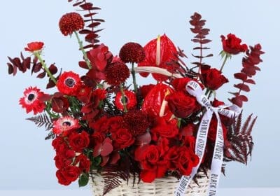 Red roses, red anthurium, red gerbera daisies, and red foliage in a wicker basket arrangement style, finished with a printed ribbon bow.