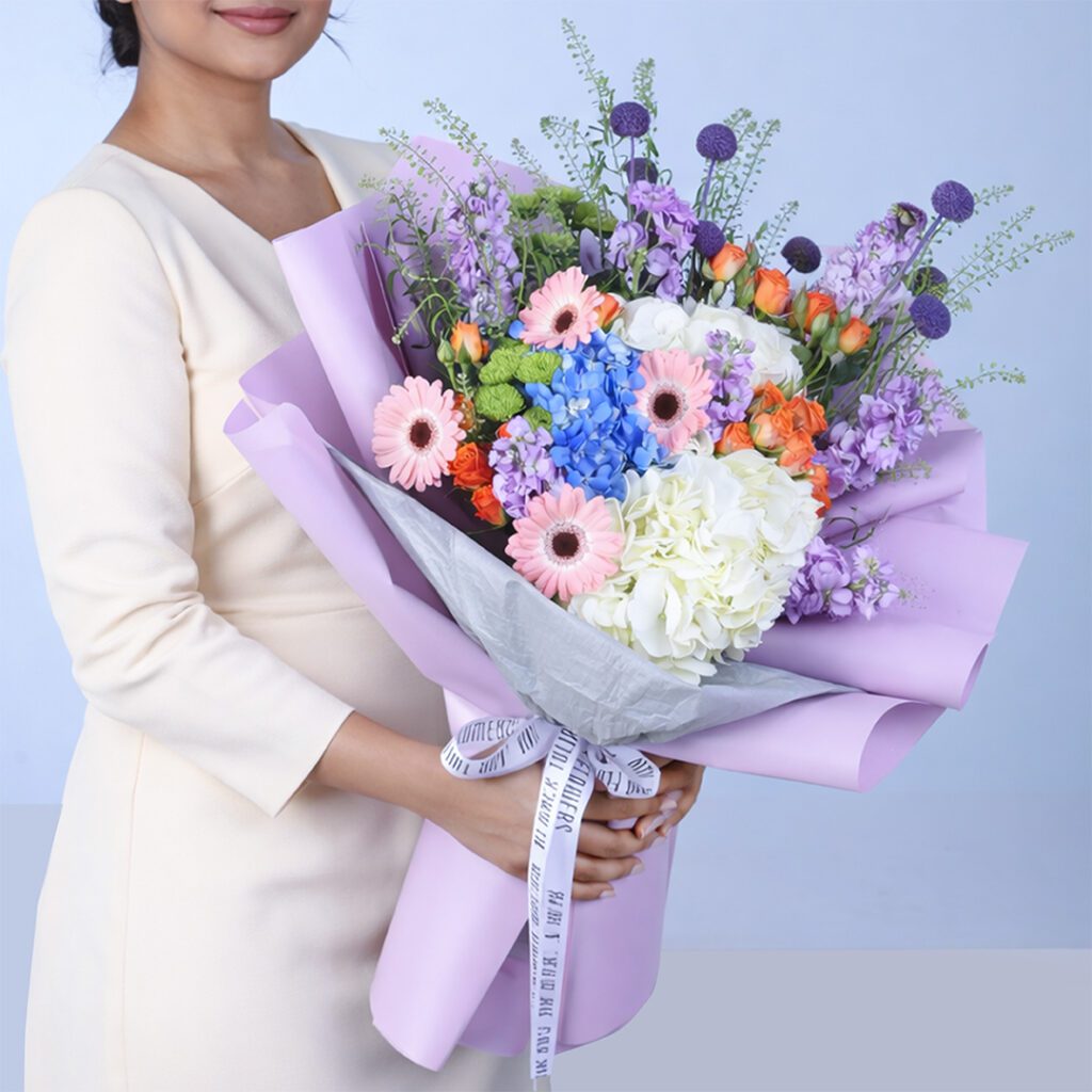 Elegant mixed pastel bouquet with pink gerberas, blue and white hydrangeas, orange roses, and purple flowers wrapped in lavender paper