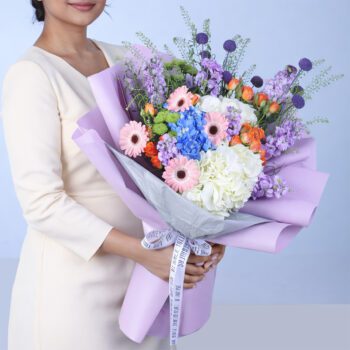 Elegant mixed pastel bouquet with pink gerberas, blue and white hydrangeas, orange roses, and purple flowers wrapped in lavender paper