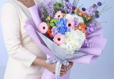 Elegant mixed pastel bouquet with pink gerberas, blue and white hydrangeas, orange roses, and purple flowers wrapped in lavender paper